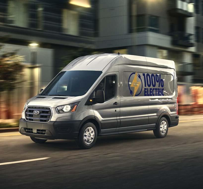 2022 Ford Transit Passenger Van traveling through the city with "100% Electric" on the side